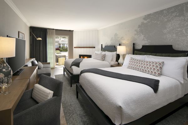 The image shows a hotel room with two double beds, a TV, a small sitting area, and a view of a patio through glass doors.