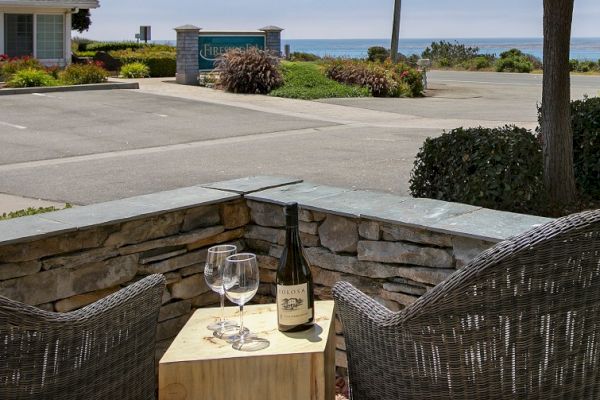 The image shows two wicker chairs, a wooden table with a wine bottle and glasses, near a stone wall overlooking a parking lot and ocean view.