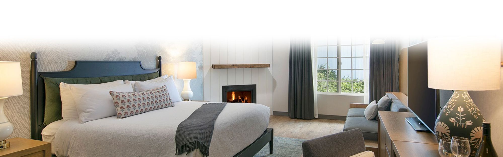 A cozy bedroom features a large bed, soft lamps, a fireplace, a window with curtains, and a TV on a dresser.