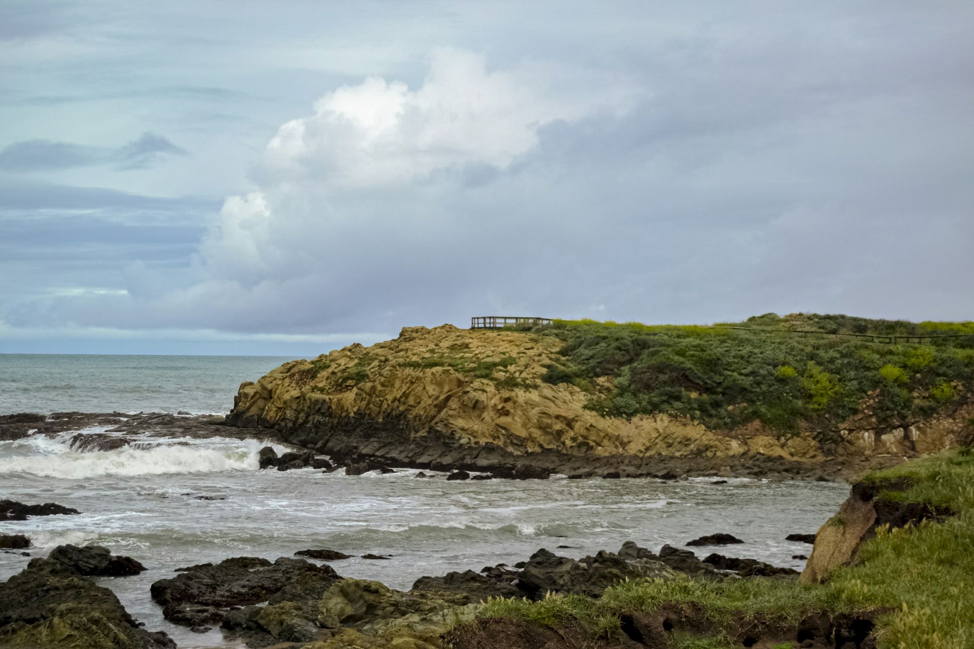 A rocky coastline with waves crashing, a grassy cliff, and dramatic clouds in the sky complete the scenic view at the shore.