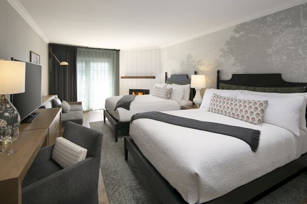 A modern hotel room with two double beds, a TV, a chair, a desk, and a balcony. It features stylish decor and soft lighting.