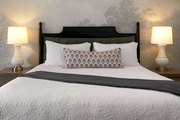 The image shows a neatly made bed with a textured quilt, decorative pillow, flanked by two side tables each with a lit lamp.