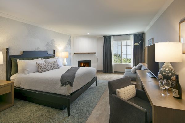 A cozy bedroom with a king-sized bed, fireplace, TV, and large window with curtains. A desk holds wine glasses and a bottle for relaxation.