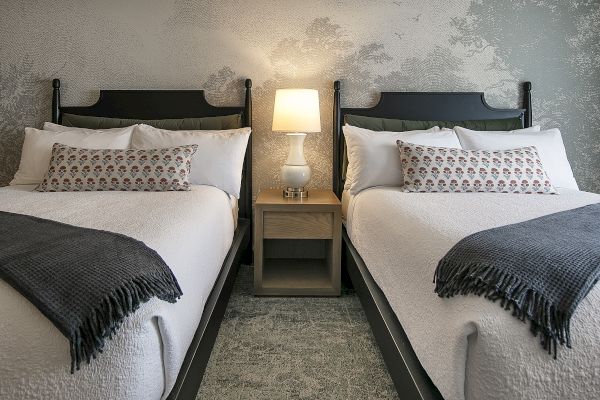 The image shows a bedroom with two neatly made beds, a nightstand with a lamp, and decorative pillows and blankets.