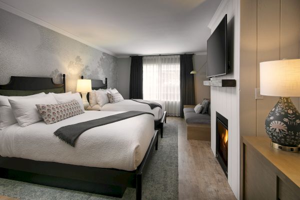 The image shows a cozy hotel room with two beds, a fireplace, a TV, and stylish decor, including lamps and cushions.