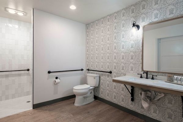 The image shows a modern bathroom with a toilet, sink, mirror, walk-in shower, and patterned wallpaper.
