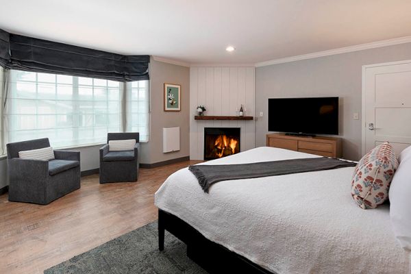 This image shows a cozy bedroom with a bed, TV, chairs, bay window, and a fireplace, creating a warm and inviting atmosphere.