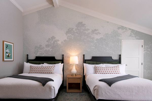 The image shows a bedroom with two beds, a nightstand with a lamp, an artistic wall design, and a small framed picture on the wall.