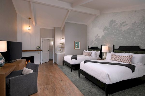 The image shows a cozy hotel room with two beds, a TV, a fireplace, and stylish decor in neutral tones, creating a comfortable atmosphere.