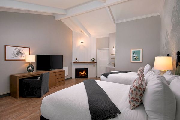 A cozy hotel room with two beds, a TV, desk, and a lit fireplace on one side, creating a warm atmosphere.