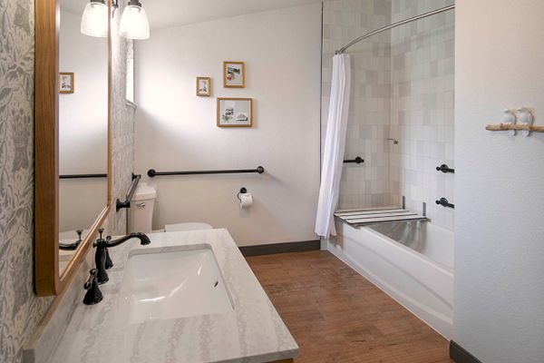 The image shows a bathroom with a double sink, a bathtub with a shower curtain, wall-mounted safety bars, and small framed pictures.