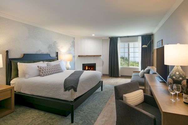 A cozy bedroom with a large bed, fireplace, desk, chairs, and lamps, featuring neutral and blue tones with a window view.