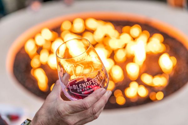A hand holding a wine glass labeled "BLENDFEST ON THE COAST" in front of a warm, glowing fire pit.