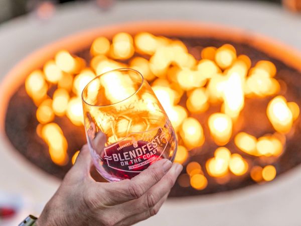 A hand holding a wine glass labeled "BLENDFEST ON THE COAST" in front of a warm, glowing fire pit.