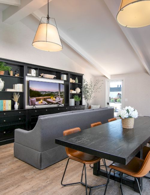 The image shows a modern living and dining area with a gray sofa, black table, brown chairs, brick fireplace, and large shelving unit.