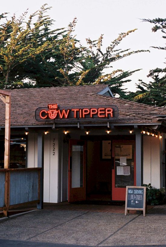 A small building with a sign that says "THE COW TIPPER" in red neon letters, surrounded by trees and some outdoor seating.