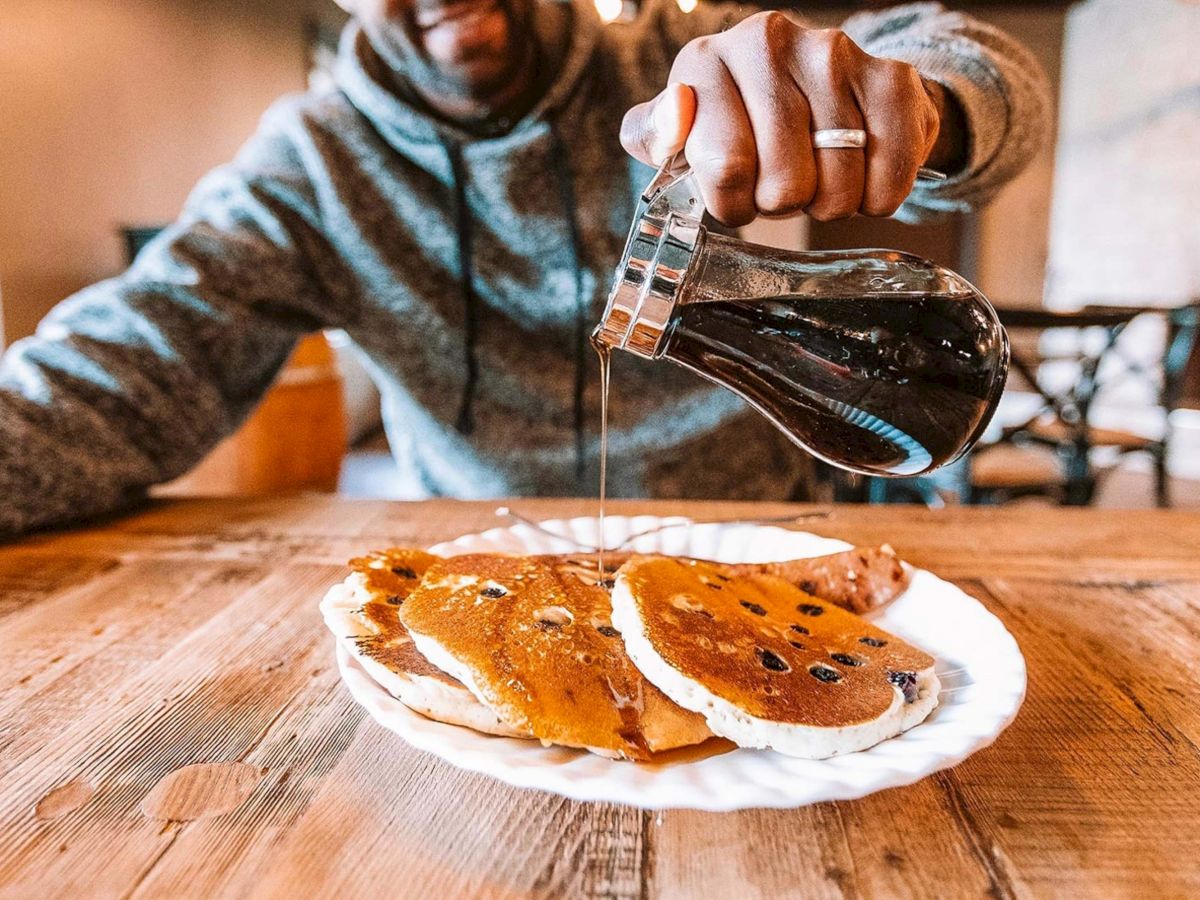A person is pouring syrup onto a stack of pancakes on a plate, set on a wooden table.