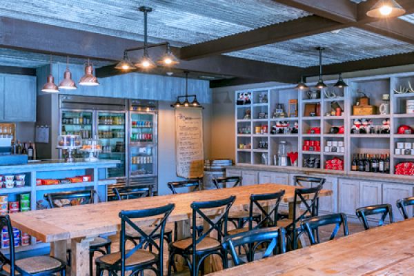 The image shows a rustic-style caf&eacute; with wooden tables, black chairs, and shelves stocked with various items. Lighting is warm and ambient.
