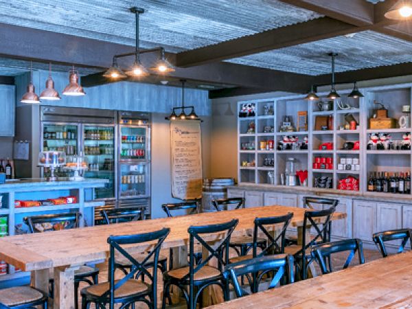 The image shows a rustic-style caf&eacute; with wooden tables, black chairs, and shelves stocked with various items. Lighting is warm and ambient.