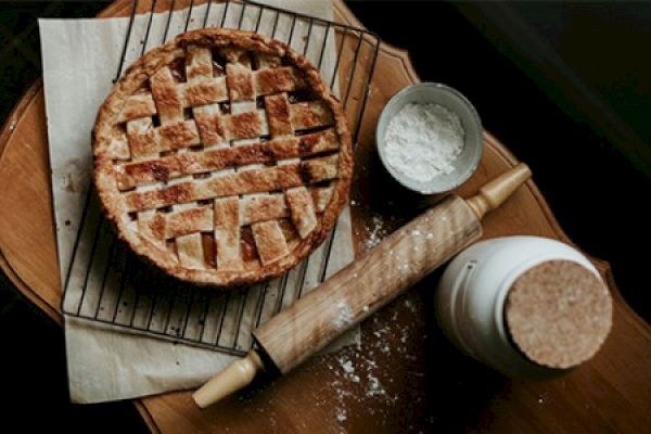 A lattice-topped pie on a cooling rack, surrounded by a rolling pin, a bowl with flour, and a jar, resting on a wooden surface.