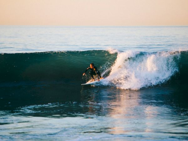 A surfer rides a green-topped wave at sunset, balanced on the board as a white crest breaks behind him, water shimmering calmly.
