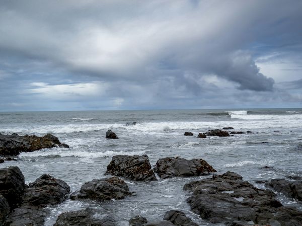 A rocky coastline with dark, jagged rocks in the foreground, blue-gray waves, and a cloudy sky above the ocean.