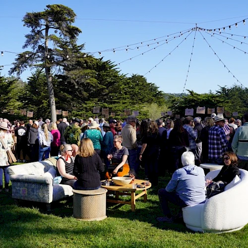 A large outdoor gathering with people seated and standing, surrounded by trees, under string lights.
