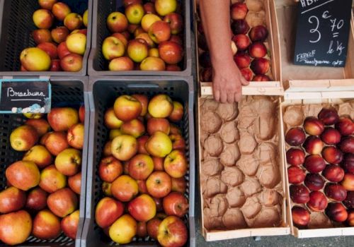 A market stall displays various apples in crates, with prices noted on chalkboards, and a person arranging them.