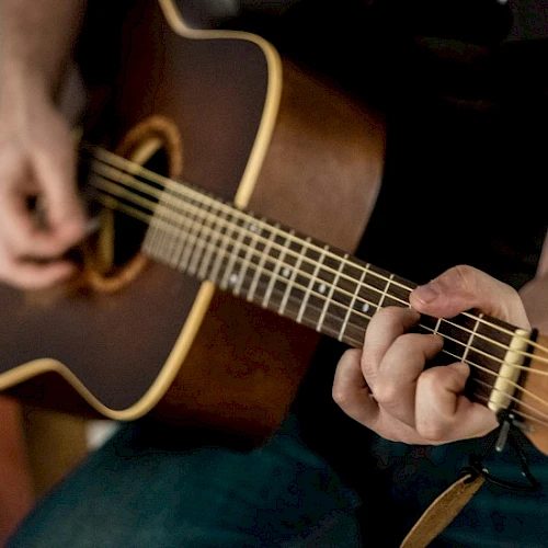A person is playing an acoustic guitar, focusing on the strings and fretboard, with hands strumming and forming chords.