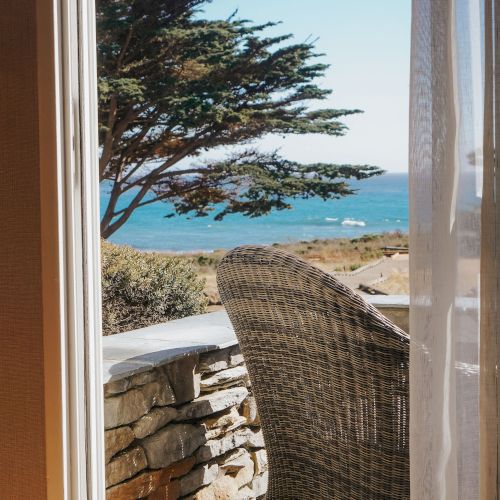 A seaside view from a doorway: a wicker chair on a stone patio, a low stone wall, a tree, and the ocean beyond, with clear blue sky.