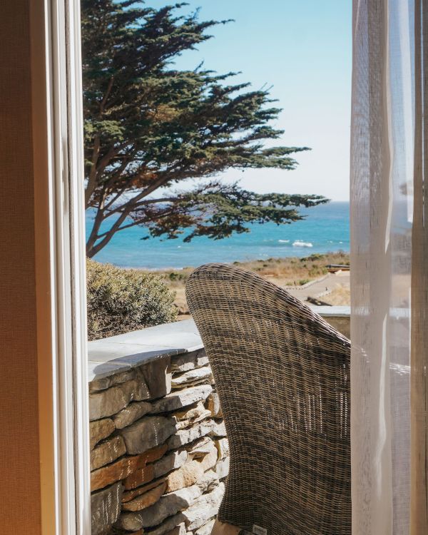 A seaside view from a doorway: a wicker chair on a stone patio, a low stone wall, a tree, and the ocean beyond, with clear blue sky.