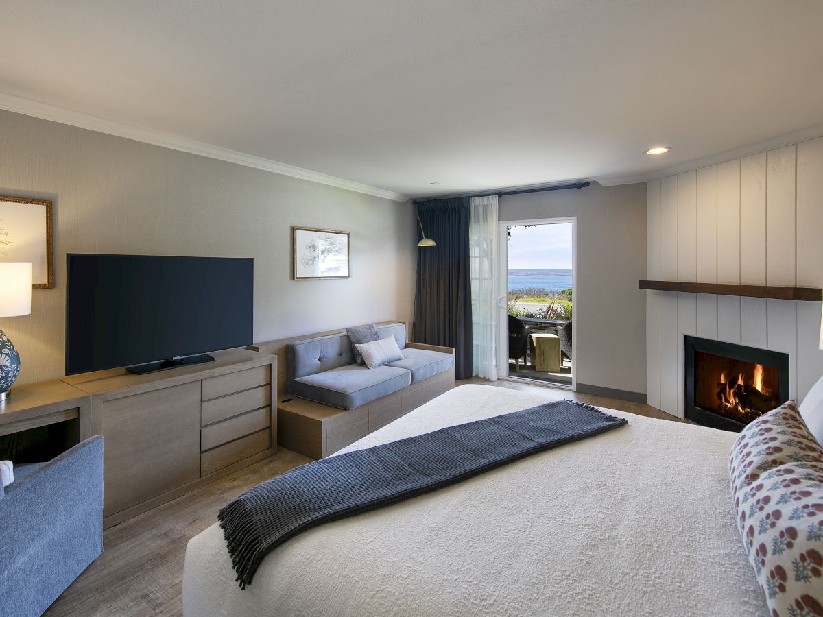 A modern hotel room with a king bed, seating area, TV, fireplace, and a balcony view, all in neutral tones.