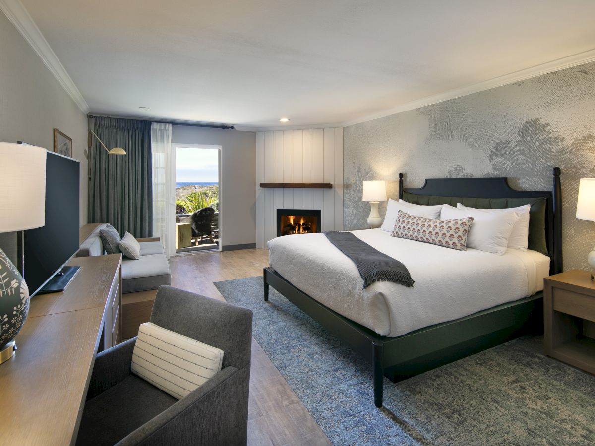 A clean, modern hotel room with a king bed, two nightstands, a fireplace, a desk and chair, a sofa, and a view to the balcony. 140 chars ending with a period.