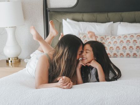 Two people whisper and cuddle on a bed, smiling as they lie close together; a cozy bedroom scene with a white bedspread and lamp nearby.