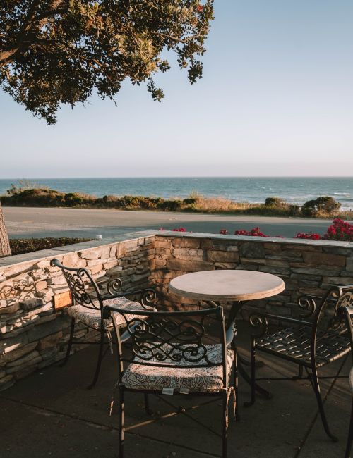 A cozy stone patio with a round table and wrought-iron chairs overlooking a calm sea and rocky shoreline, framed by a leafy tree branch.