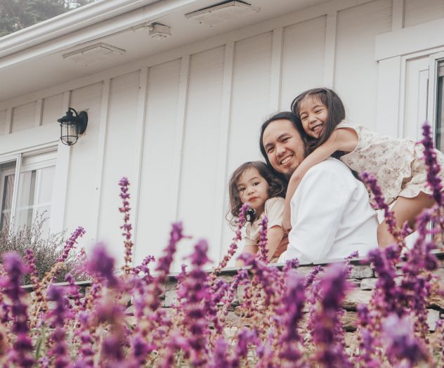A family hug on a porch, smiling together behind purple flowers in bloom under a white house.