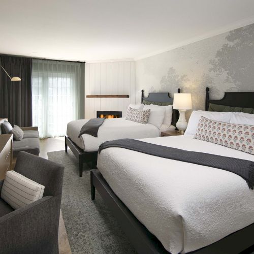 A modern hotel room with two large beds, clean white bedding and gray accents, a desk with chair, a TV, and a seating area near a window.