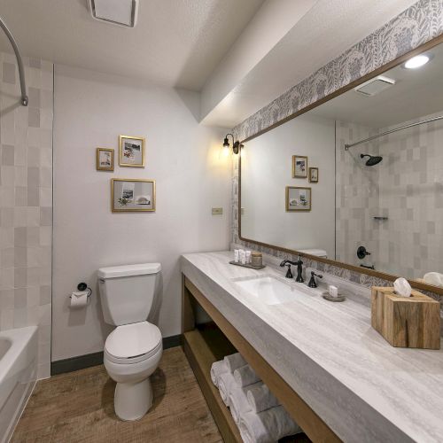 A modern bathroom with a long marble counter, double sink, large framed mirror, a white toilet, and a tub/shower combo with tiled walls.