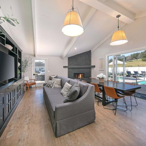 A bright, modern open-plan living space with a cozy gray sofa, glass coffee table, warm wood flooring, pendant lights, and a stylish kitchen in the background.