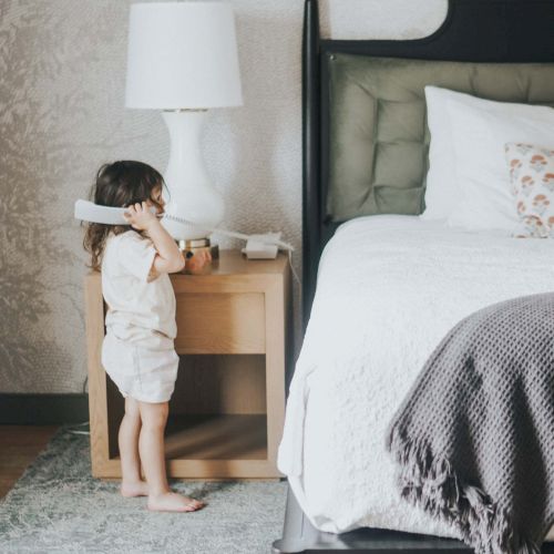 A child stands by a dresser in a light-filled bedroom, near a neatly made bed with a gray and white color scheme.