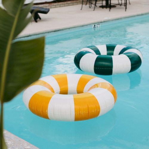 A pool with a few colorful inner tubes (orange, white, and teal) floating near the edge, lounge chairs in the background, and palm leaves.