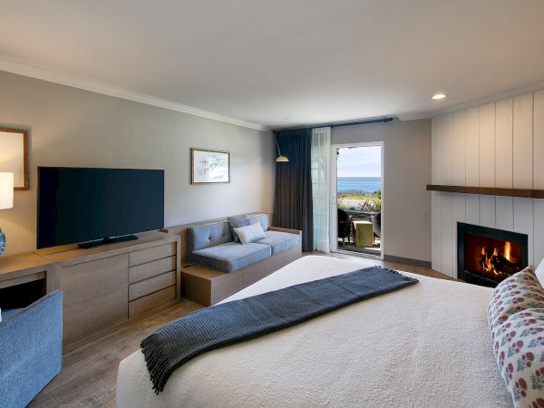 A modern hotel room with a large bed, a flat-screen TV on a dresser, a sitting area, fireplace, balcony view, and soft neutral decor, ending here.