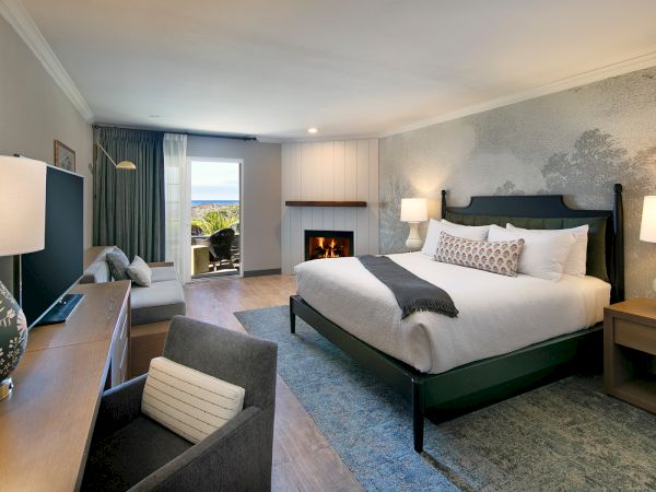 A stylish hotel room featuring a king bed with throw blanket, nightstands, lamps, a desk and chair, a seating area, a fireplace, and a view toward a balcony or window.