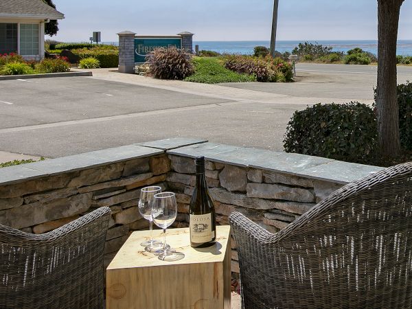 A cozy outdoor setup with two wicker chairs facing a small wooden table, a wine bottle and two glasses, overlooking a coastal road and blue ocean.
