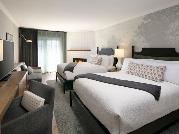 Two large beds in a bright hotel room with a desk, seating area, lamps, and a soft, neutral decor.