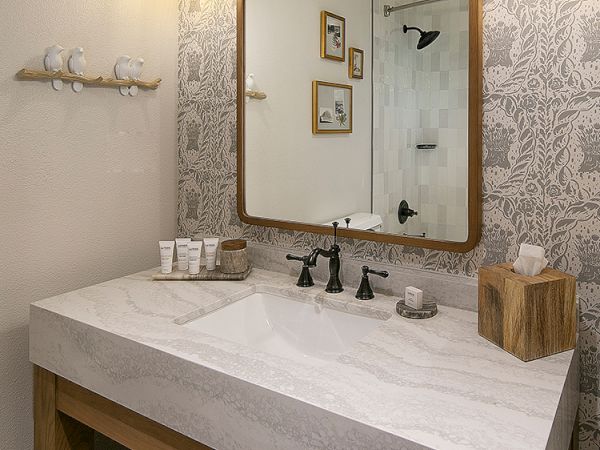 A modern bathroom vanity with a marble sink, wooden frame mirror, patterned wallpaper, wall sconces, and neatly arranged toiletries and towels.