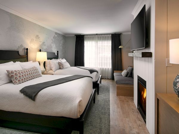 A stylish hotel room with two queen beds, neutral decor, nightstands, a fireplace, a wall-mounted TV, and a sitting area by a window.