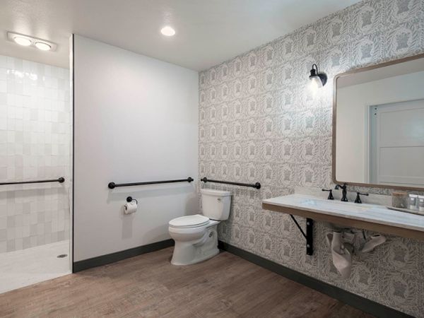 A spacious, accessible bathroom with a toilet, a wall-mounted sink, a large mirror, and patterned wallpaper; includes grab bars and a partial shower area.