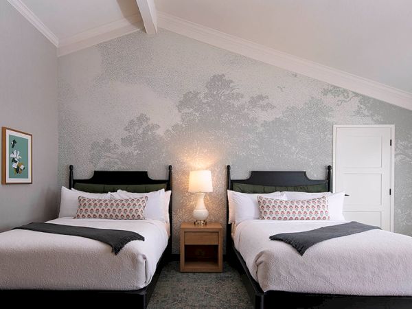 Two twin beds with dark wood headboards, a shared nightstand, lamp, soft gray walls with subtle mural, and a closed white door in a tidy hotel room.