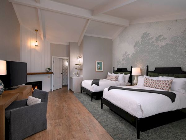 A modern hotel room with two queen beds, gray walls with a textured accent, a seating area, a desk, and warm lighting in a clean, airy space.